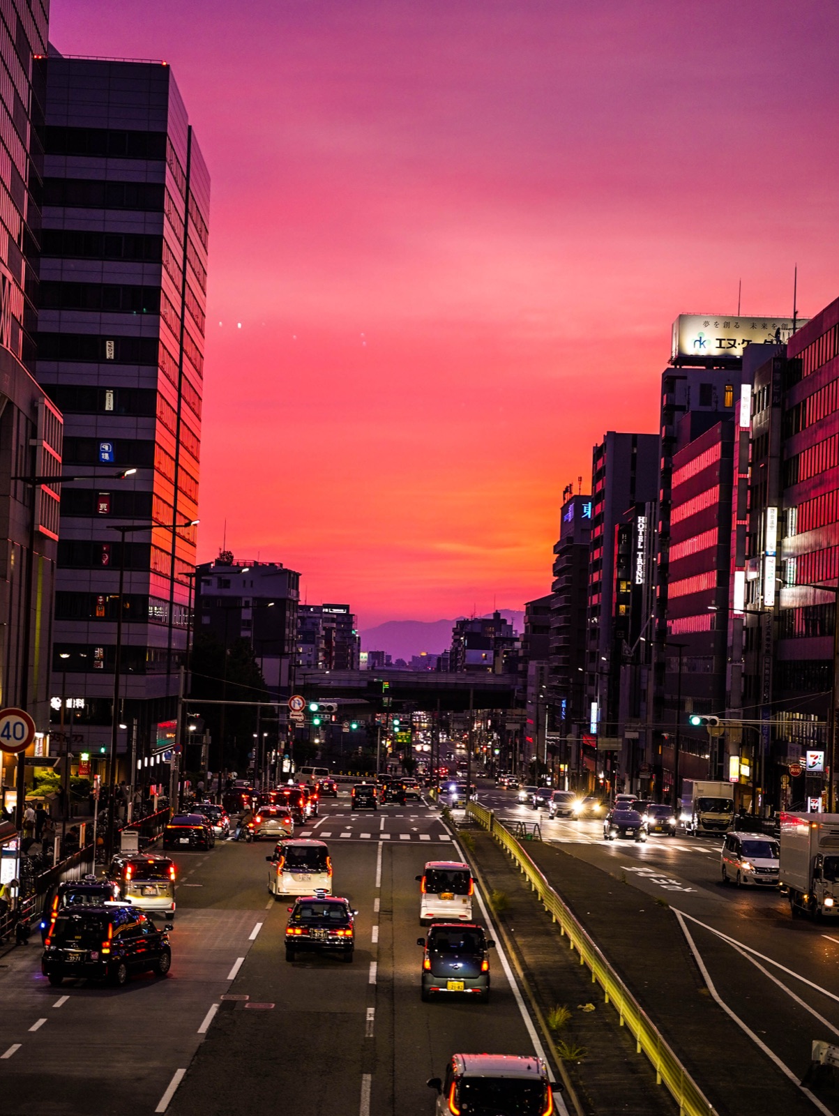 Traffic fills a city avenue beneath a rose colored evening sky.
