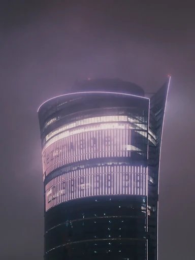 A curved glass tower glows through a misty night sky.