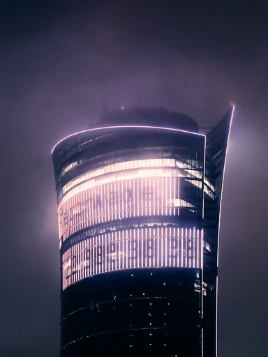 A curved glass tower glows through a misty night sky.