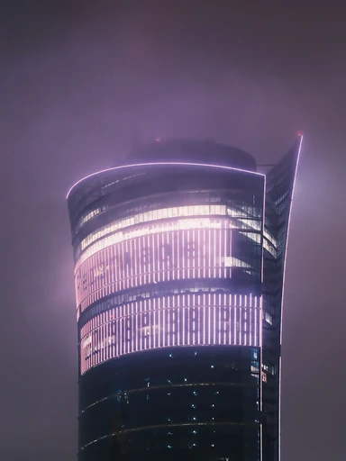 A curved glass tower glows through a misty night sky.