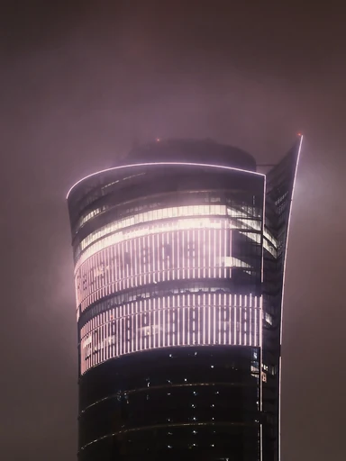 A curved glass tower glows through a misty night sky.