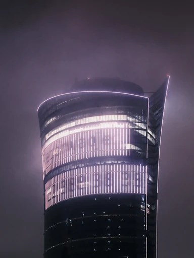 A curved glass tower glows through a misty night sky.