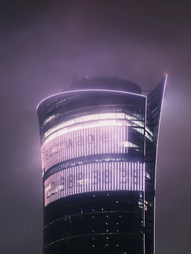 A curved glass tower glows through a misty night sky.