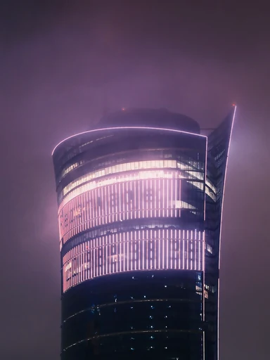 A curved glass tower glows through a misty night sky.