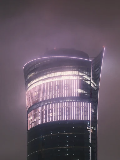A curved glass tower glows through a misty night sky.