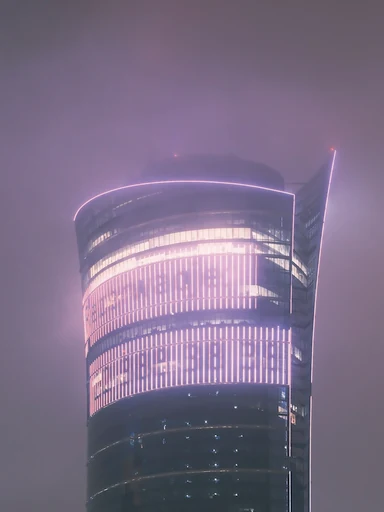 A curved glass tower glows through a misty night sky.